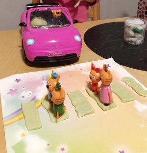 Kotki przechodzące przez jezdnię wykonaną z plasteliny, w tle kotek w różowym samochodzie