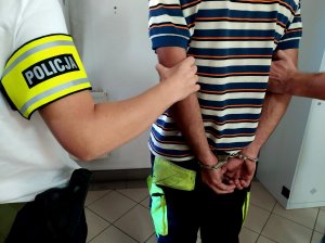 mężczyzna w kajdankach założonych na ręce trzymane z tyłu policjanci trzymający go za ręce